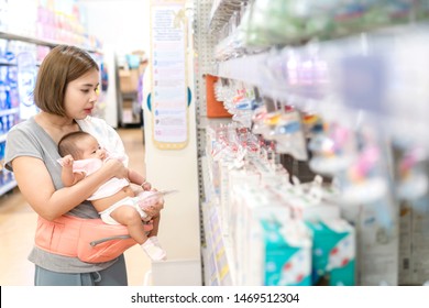 Young Asian Mother And Little Cute Baby In Carrier Are Shopping Newborn Baby Product In Supermarket. Mom Is Choosing New Nipple For Her Child.