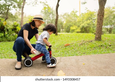 Young Asian Mother Teaching Her Adorable Toddler Girl To Ride The Bike For The First Time While Spending Time Outing Together In The Morning, Concept Mother And Child Love, Family Time And Activity.