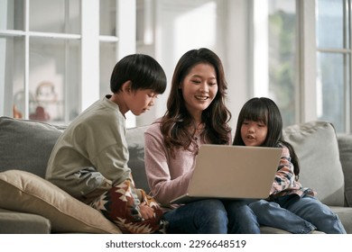 young asian mother and two children sitting on family couch at home using laptop computer together - Shutterstock ID 2296648519