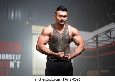 Young Asian muscular male fitness trainer in a tank top standing with hold his arms down, flexing arm and shoulder muscles while looking forward with a serious face with a blurred background of gym. - Powered by Shutterstock