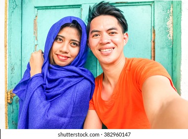 Young Asian Muslim Couple Taking Selfie Next To Old Green Wooden Door - Cheerful Teenagers Of Different Religion Posing For Self Photo - Portrait Of Mixed Culture Students - Focus On Male Subject