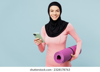 Young asian muslim fitness trainer sporty woman wear pink abaya hijab spend time in home gym hold yoga mat use mobile cell phone isolated on plain blue background studio. Workout sport fit abs concept - Powered by Shutterstock