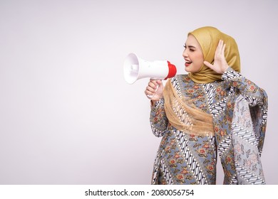 Young Asian Muslim Girl In Batik Dress  With Hijab Holds In Hand Bullhorn Public Address Loud Hailer Isolated On White Studio Background.  Scream In Megaphone. People Emotions Lifestyle Concept.