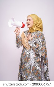 Young Asian Muslim Girl In Batik Dress  With Hijab Holds In Hand Bullhorn Public Address Loud Hailer Isolated On White Studio Background.  Scream In Megaphone. People Emotions Lifestyle Concept.