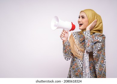 Young Asian Muslim Girl In Batik Dress  With Hijab Holds In Hand Bullhorn Public Address Loud Hailer Isolated On White Studio Background.  Scream In Megaphone. People Emotions Lifestyle Concept.