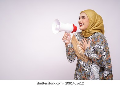 Young Asian Muslim Girl In Batik Dress  With Hijab Holds In Hand Bullhorn Public Address Loud Hailer Isolated On White Studio Background.  Scream In Megaphone. People Emotions Lifestyle Concept.