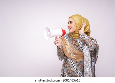 Young Asian Muslim Girl In Batik Dress  With Hijab Holds In Hand Bullhorn Public Address Loud Hailer Isolated On White Studio Background.  Scream In Megaphone. People Emotions Lifestyle Concept.