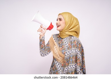 Young Asian Muslim Girl In Batik Dress  With Hijab Holds In Hand Bullhorn Public Address Loud Hailer Isolated On White Studio Background.  Scream In Megaphone. People Emotions Lifestyle Concept.