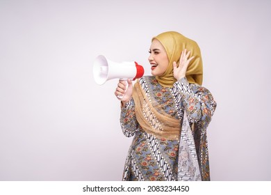 Young Asian Muslim Girl In Batik Dress  With Hijab Holds In Hand Bullhorn Public Address Loud Hailer Isolated On White Studio Background.  Scream In Megaphone. People Emotions Lifestyle Concept.