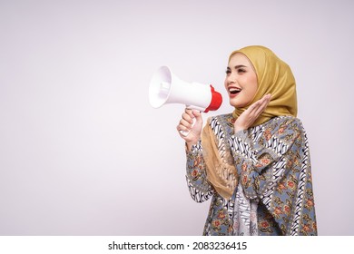 Young Asian Muslim Girl In Batik Dress  With Hijab Holds In Hand Bullhorn Public Address Loud Hailer Isolated On White Studio Background.  Scream In Megaphone. People Emotions Lifestyle Concept.