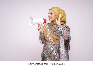 Young Asian Muslim Girl In Batik Dress  With Hijab Holds In Hand Bullhorn Public Address Loud Hailer Isolated On White Studio Background.  Scream In Megaphone. People Emotions Lifestyle Concept.