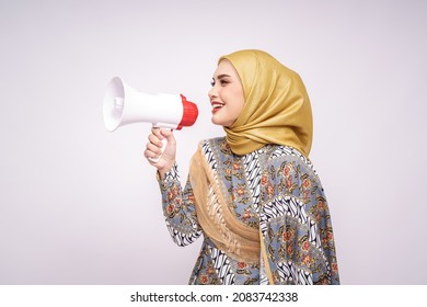 Young Asian Muslim Girl In Batik Dress  With Hijab Holds In Hand Bullhorn Public Address Loud Hailer Isolated On White Studio Background.  Scream In Megaphone. People Emotions Lifestyle Concept.