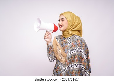 Young Asian Muslim Girl In Batik Dress  With Hijab Holds In Hand Bullhorn Public Address Loud Hailer Isolated On White Studio Background.  Scream In Megaphone. People Emotions Lifestyle Concept.