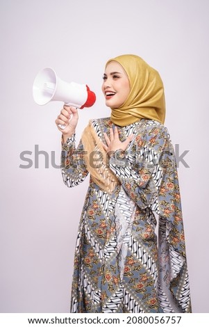 Young Asian muslim girl in batik dress  with hijab holds in hand bullhorn public address loud hailer isolated on white studio background.  Scream in megaphone. People emotions lifestyle concept.