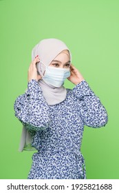 A Young Asian Muslim Woman With Hijab Wearing A Surgical Mask Isolated Over Green Background Studio. Coronavirus Covid-19 Prevention Concept.