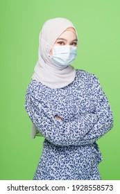 A Young Asian Muslim Woman With Hijab Wearing A Surgical Mask Isolated Over Green Background Studio. Coronavirus Covid-19 Prevention Concept.