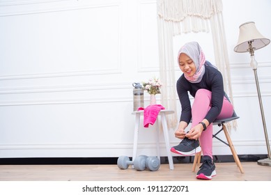 Young Asian Muslim Woman In Pink Workout Dress Tying Her Shoelaces In Her Living Room, Preparing For Workout, Indoor Home Exercise Concept