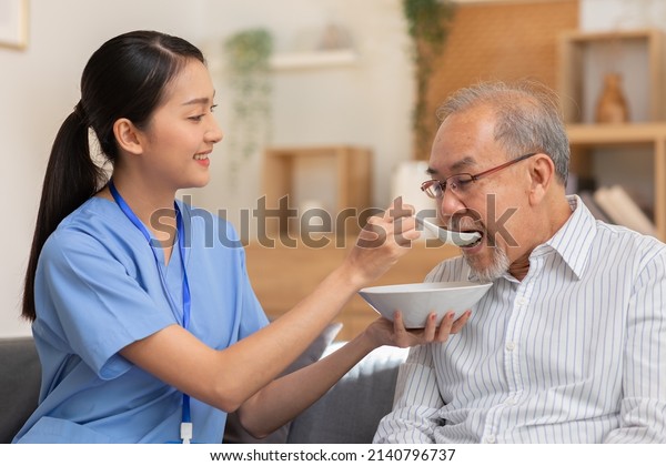 Young Asian nurse feeding breakfast to senior man\
at couch. Old Asian man with white beard enjoy his meal. Caregiver\
take care of senior man, give soft food, look after to\
patient.Elderly Care\
Concept