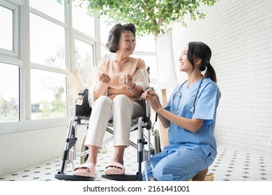 Young Asian Nurse Talking To A Smiling Senior Female Patient On A Wheelchair And Comforting Her.