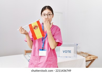 Young Asian Nurse Woman At Political Campaign Election Holding Spain Flag Covering Mouth With Hand, Shocked And Afraid For Mistake. Surprised Expression 