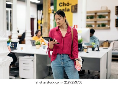 Young Asian Office Worker Standing In The Office Using Her Digital Tablet Holding A Water Bottle.