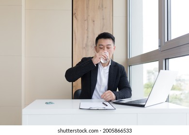 Young Asian Office Worker Suffering From Headache At Work In The Office Drinks Medicine Pill With Water. Sick Man In A Suit At The Computer Indoors With Severe Pain Ache Uses The Drug In Workplace