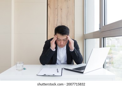 Young Asian Office Worker Suffering From Headache At Work In The Office Drinks Medicine Pill With Water. Sick Man In A Suit At The Computer Indoors With Severe Pain Ache Uses The Drug In Workplace