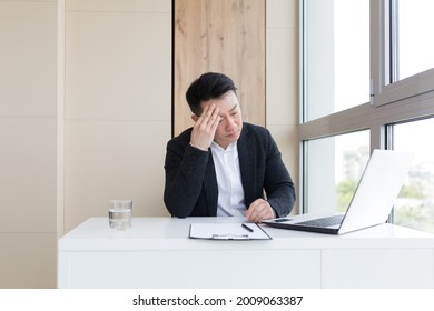 Young Asian Office Worker Suffering From Headache At Work In The Office Drinks Medicine Pill With Water. Sick Man In A Suit At The Computer Indoors With Severe Pain Ache Uses The Drug In Workplace