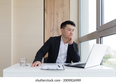 Young Asian Office Worker Suffering From Headache At Work In The Office Drinks Medicine Pill With Water. Sick Man In A Suit At The Computer Indoors With Severe Pain Ache Uses The Drug In Workplace