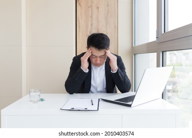 Young Asian Office Worker Suffering From Headache At Work In The Office Drinks Medicine Pill With Water. Sick Man In A Suit At The Computer Indoors With Severe Pain Ache Uses The Drug In Workplace