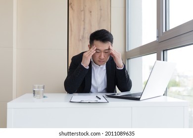 Young Asian Office Worker Suffering From Headache At Work In The Office Drinks Medicine Pill With Water. Sick Man In A Suit At The Computer Indoors With Severe Pain Ache Uses The Drug In Workplace