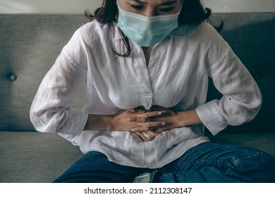 Young Asian Patient Woman Wearing A Face Mask Suffering From Abdominal Pain, Period, Or Menstrual While Sitting On A Sofa At Home Or Hospital. Health Care And Health Women Problem Concept.