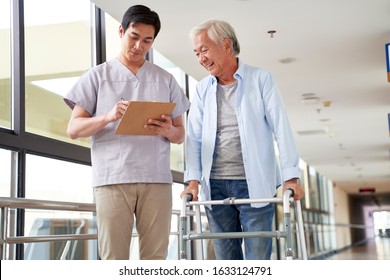 Young Asian Physical Therapist Working With Old Man On Working Using A Walker In Hallway Of Nursing Home