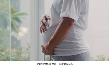 Young Asian Pregnant woman holding her belly talking with her child. Mom feeling happy smiling positive and peaceful while take care baby, pregnancy near window in living room at home concept.