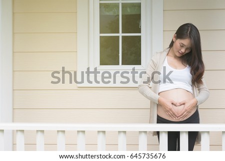 young asian pregnant woman making heart shape sign on her tummy in front of her house.  pregnancy, maternity belly care concept