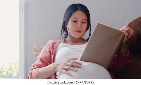 Young Asian Pregnant Woman Read A Book For Baby In Belly. Mom Feeling Happy Smiling Positive And Peaceful While Take Care Child Lying On Sofa In Living Room At Home Concept.