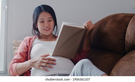 Young Asian Pregnant Woman Read A Book For Baby In Belly. Mom Feeling Happy Smiling Positive And Peaceful While Take Care Child Lying On Sofa In Living Room At Home Concept.