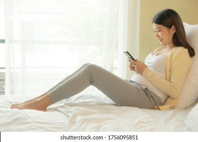 Young Asian Pregnant Woman Using Mobile Phone In Bedroom.
Japanese Girl Use Cellphone On Bed At Home.
Happy Young Pregnant Woman Sitting On Bed Looking At Smartphone Screen.