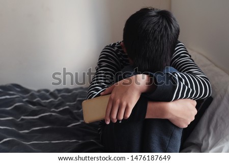 Young Asian preteen teenager boy hugging his knee in his bedroom with smartphone, Cyber bullying in kid, depressed child mental illness health, fear of missing out, FOMO