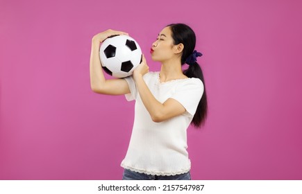 Young asian pretty girl in white t-shirt holding and kissing a football ball on the pink screen background.
