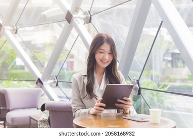 Young Asian Professional Business Woman Is Looking At Tablet Or Notepad In Her Hands While She Is Sitting At Coffee Shop Among Business Background In The City.