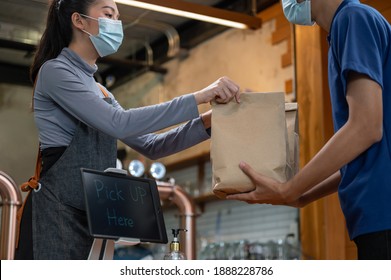 Young Asian Restaurant Owner Or Waitress Wearing Face Mask Giving Takeaway Food Bag To Delivery Man. Male Courier Taking Online Order For Delivery At Restaurant. Takeaway Food During Covid-19 Pandemic