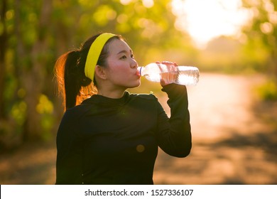 Young Asian Runner Girl Drinking Water Sweaty And Tired. Attractive And Exhausted Korean Woman Thirsty After Running Workout And Jogging At Beautiful Park On Sunset In Fitness Lifestyle