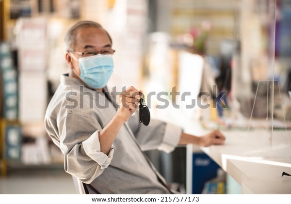 Young Asian sales man wearing covid-19\
face protection mask while working on laptop and sitting in garage\
giving car keys to senior customer during\
pandemic