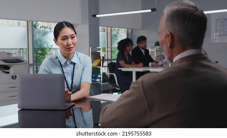 Young Asian Saleswoman With Laptop Advise Mature Client. Senior Businessman Consult Bank Employee About Loan For Startup Business