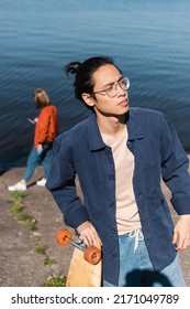 Young Asian Skater In Blue Shirt And Eyeglasses Looking Away On Riverside