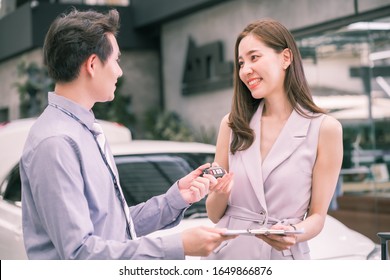 Young Asian Smiling Woman Getting Keys Of A New Car. Professional Salesperson During Work With Customer At Car Dealership. Giving Keys To New Car Owner.Concept For Car Rental Or Sale. 