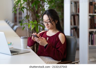 Young asian student girl procrastinate in library using smartphone and chatting in social media while preparing for exam test. Lazy woman with mobile phone in hand avoid learning and doing homework