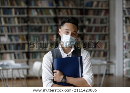 Young Asian student wearing face mask, posing in university library. Young college guy staying safe from viral infection, flu, coronavirus disease, looking at camera, smiling. Head shot portrait