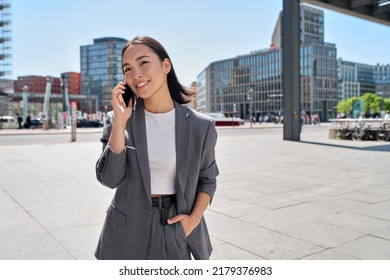 Young Asian successful businesswoman leader wearing suit standing in big city talking on mobile phone. Smiling woman making business call on cell walking on busy downtown street outdoors. - Powered by Shutterstock